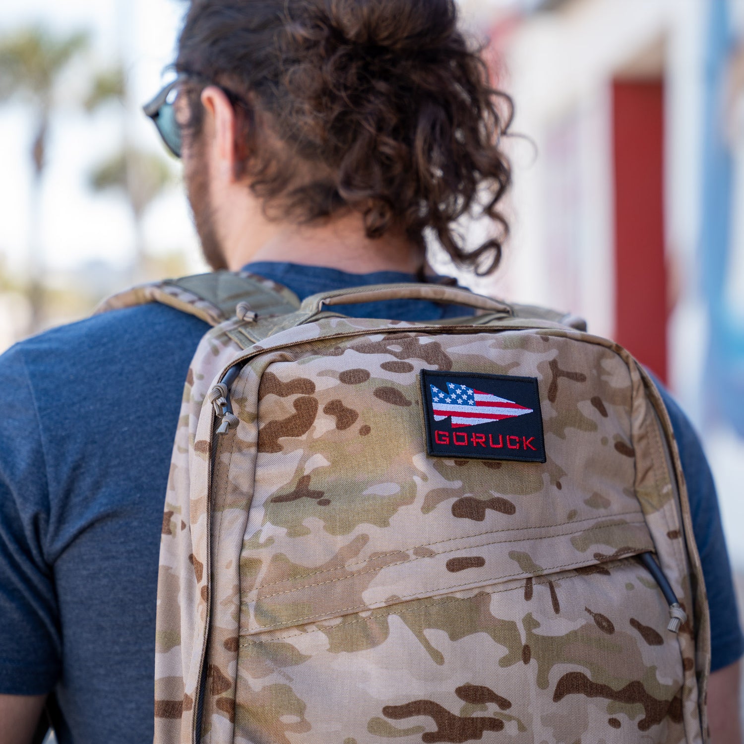 Person with long hair, seen from behind, wears a camo GR1 USA - Cordura rucksack—an everyday carry backpack made to Special Forces standards—with an American flag patch.