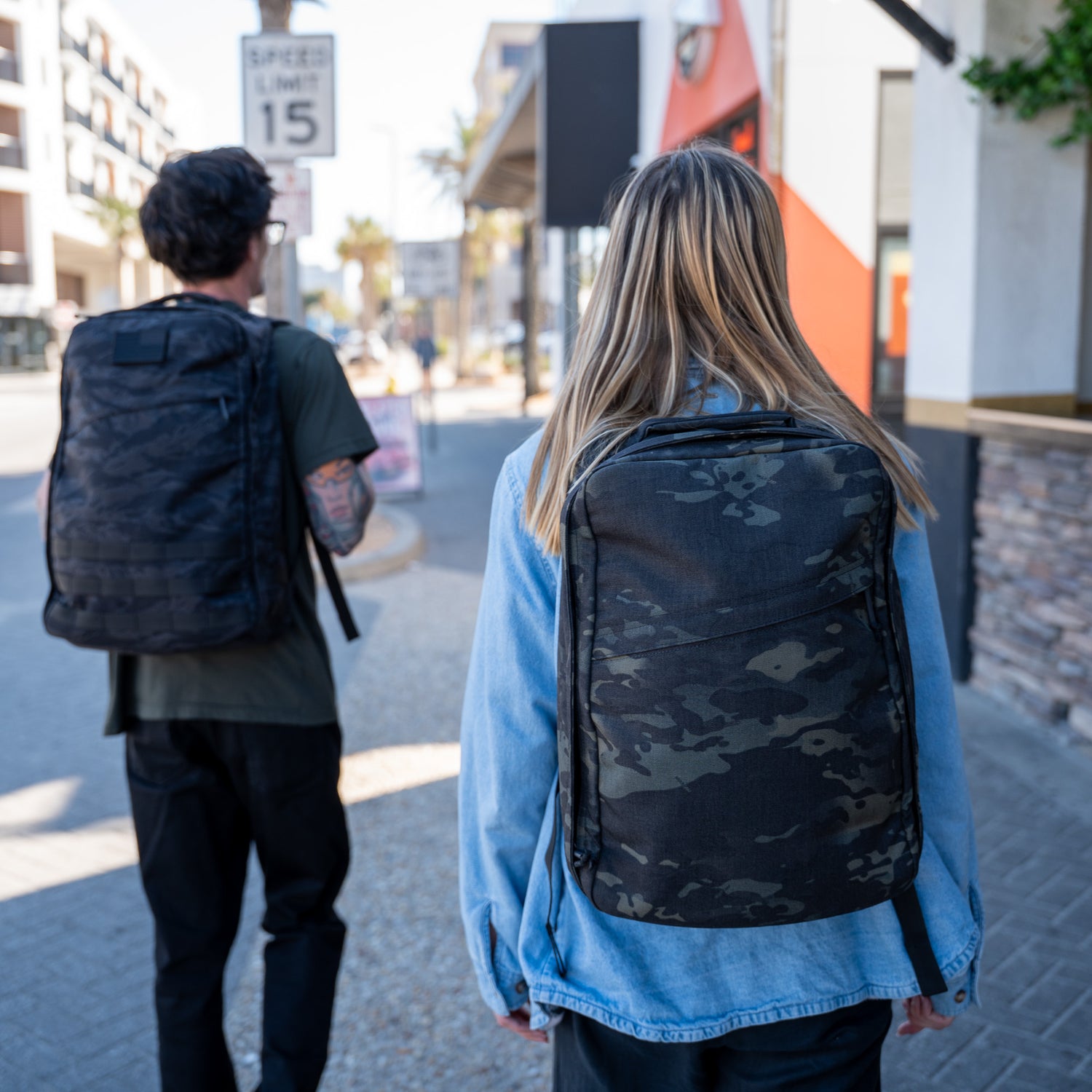Two people with GR1 USA Slick - Ballistic Nylon Cordura backpacks walk down a sunny city sidewalk—their tough, American-made gear features bombproof laptop compartments.