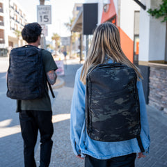 Two people with GR1 USA Slick - Ballistic Nylon Cordura backpacks walk down a sunny city sidewalk—their tough, American-made gear features bombproof laptop compartments.