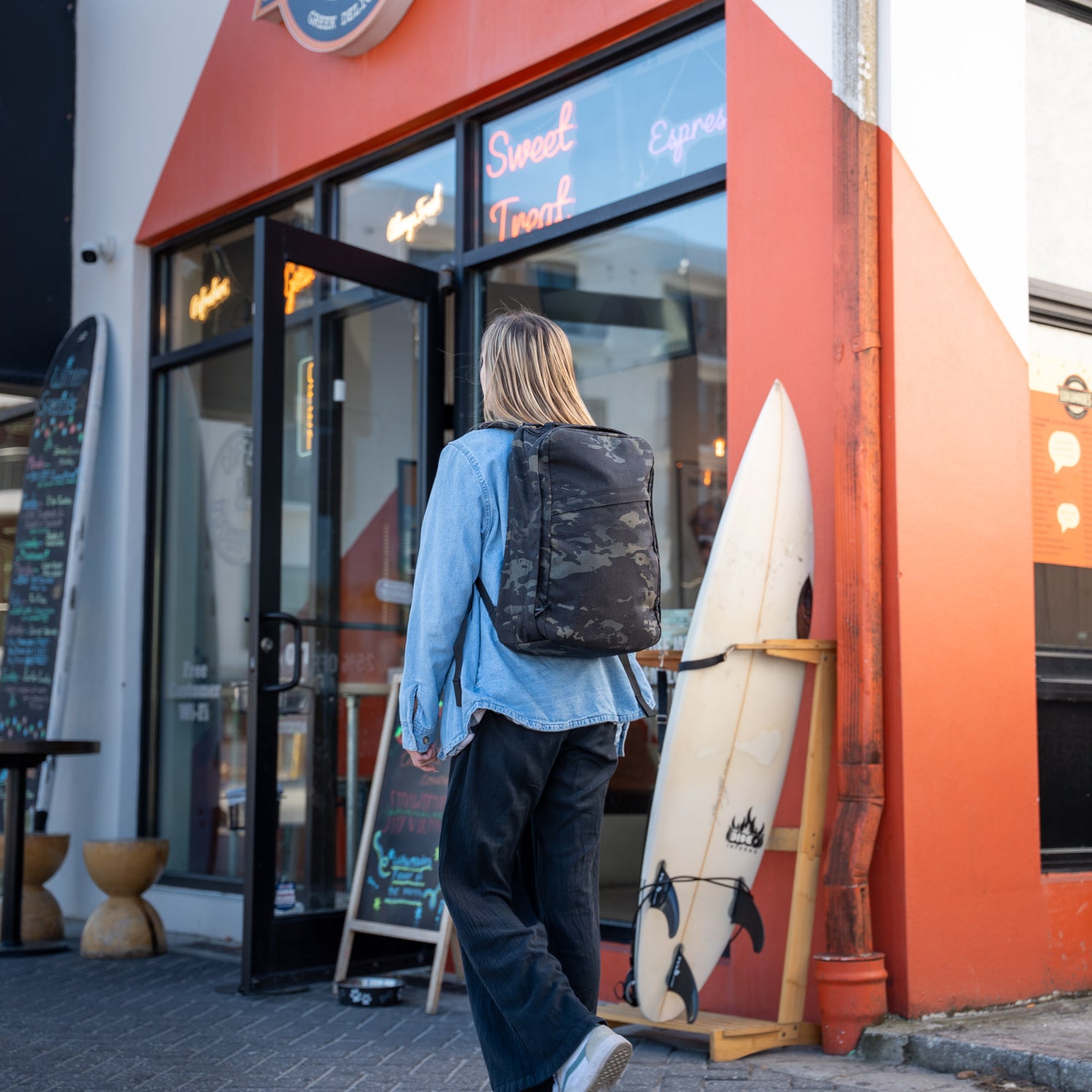 A person carrying a GR1 USA Slick - Ballistic Nylon Cordura backpack, famous for its bombproof laptop compartment and American craftsmanship, walks into a shop with a surfboard outside and an orange triangular entrance.