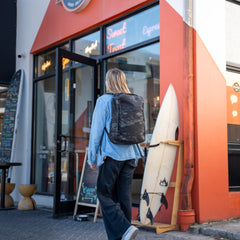 A person carrying a GR1 USA Slick - Ballistic Nylon Cordura backpack, famous for its bombproof laptop compartment and American craftsmanship, walks into a shop with a surfboard outside and an orange triangular entrance.