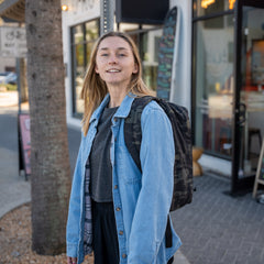 Young woman in a denim jacket with a GR1 USA Slick backpack made from Ballistic Nylon Cordura, featuring a bombproof laptop compartment, stands on a city sidewalk and smiles at the camera.