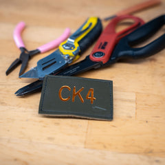 A Patch - Custom Leather Monogram rests on a wooden surface with pliers and utility knives in the background, ideal for showcasing your custom initials or gifting a personalized touch.