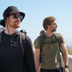 Two men wearing Ruck Plate Carrier 3.0 backpacks with ergonomic lumbar support walk outdoors on a sunny day, with others in the background.