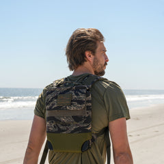 A man wearing the Ruck Plate Carrier 3.0 with ergonomic lumbar support walks along a sandy ocean beach beneath a clear blue sky.