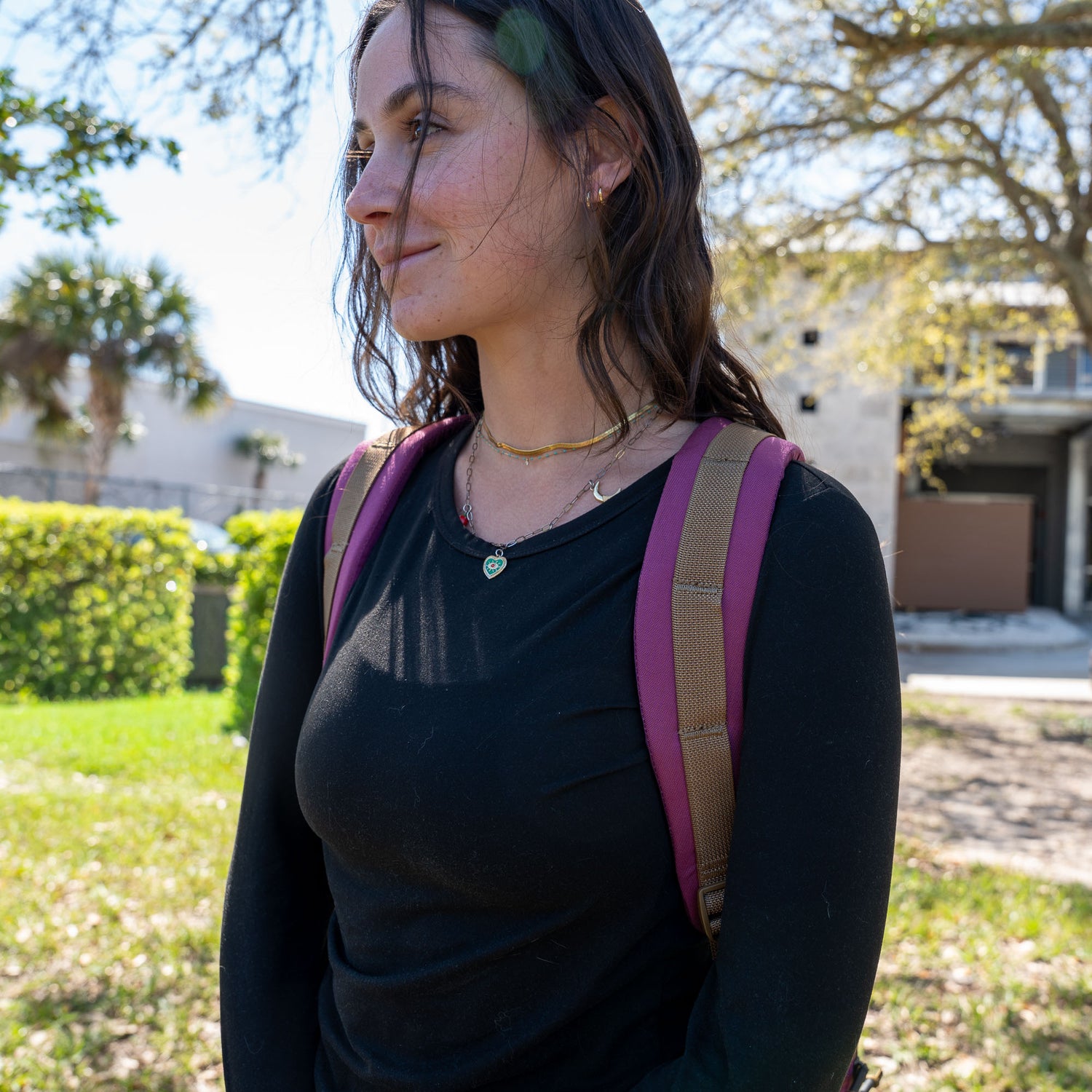 A young woman with long brown hair and a pink Bullet Ruck Classic - Ballistic Nylon Cordura 15L daypack stands outside on a sunny day, smiling slightly.