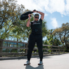 A man in athletic wear lifts a Bulgarian Sandbag with reinforced handles overhead outdoors on a sunny day, with trees and blue sky in the background.
