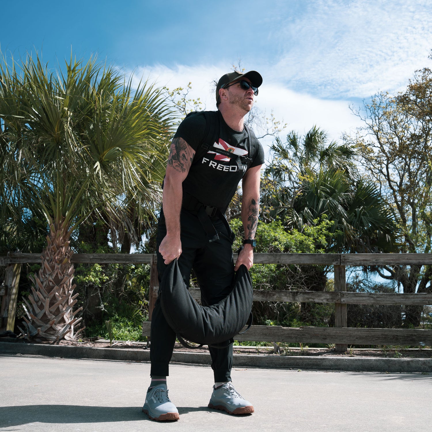 A man lifts a Bulgarian Sandbag with reinforced handles outdoors, framed by palm trees and a wooden fence in the background, while wearing sunglasses.