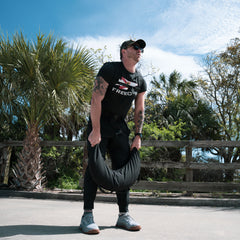 A man lifts a Bulgarian Sandbag with reinforced handles outdoors, framed by palm trees and a wooden fence in the background, while wearing sunglasses.