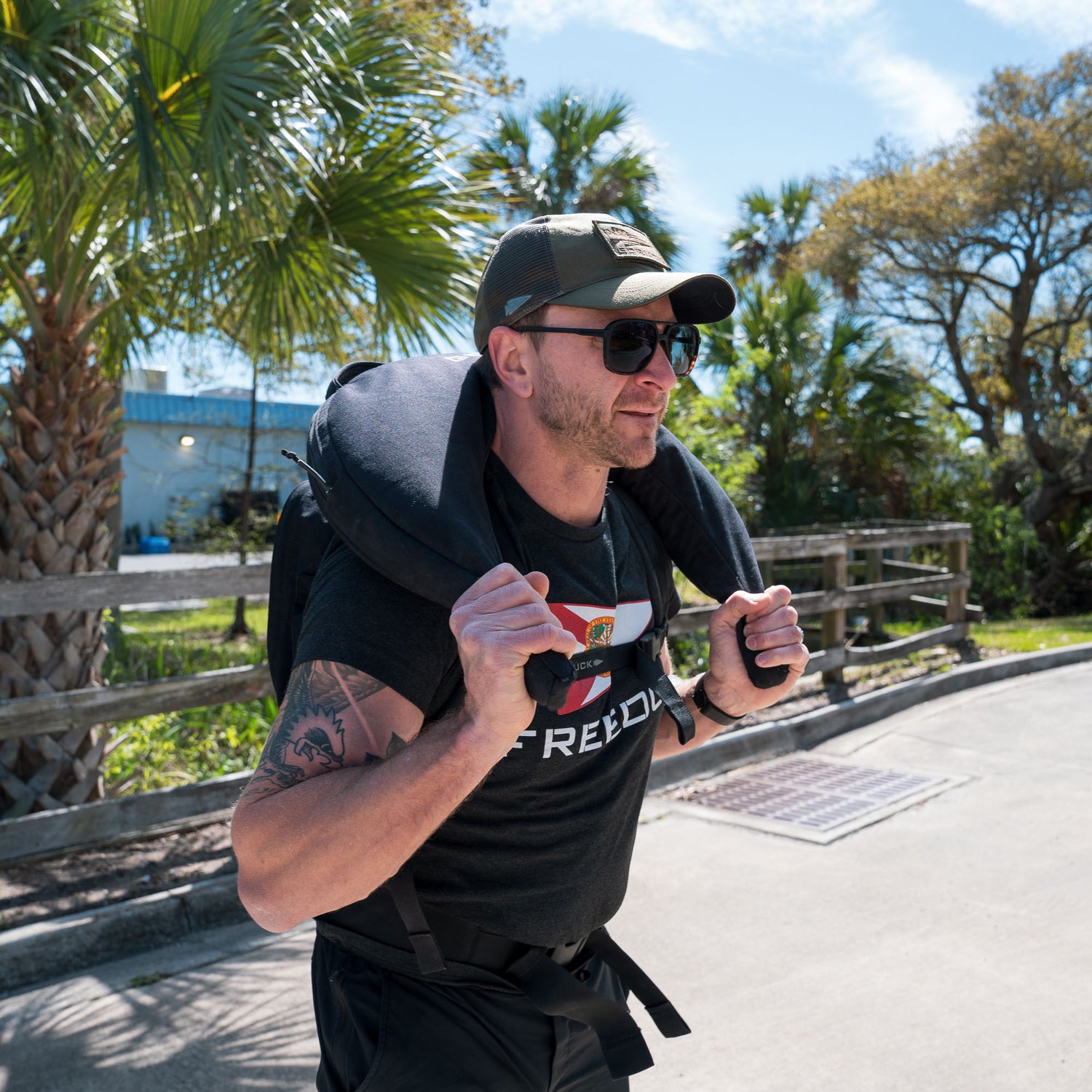 A man in sunglasses and a cap carries the Bulgarian Sandbag on his shoulders outdoors, surrounded by palm trees—the ideal scene to highlight this versatile fitness training equipment.