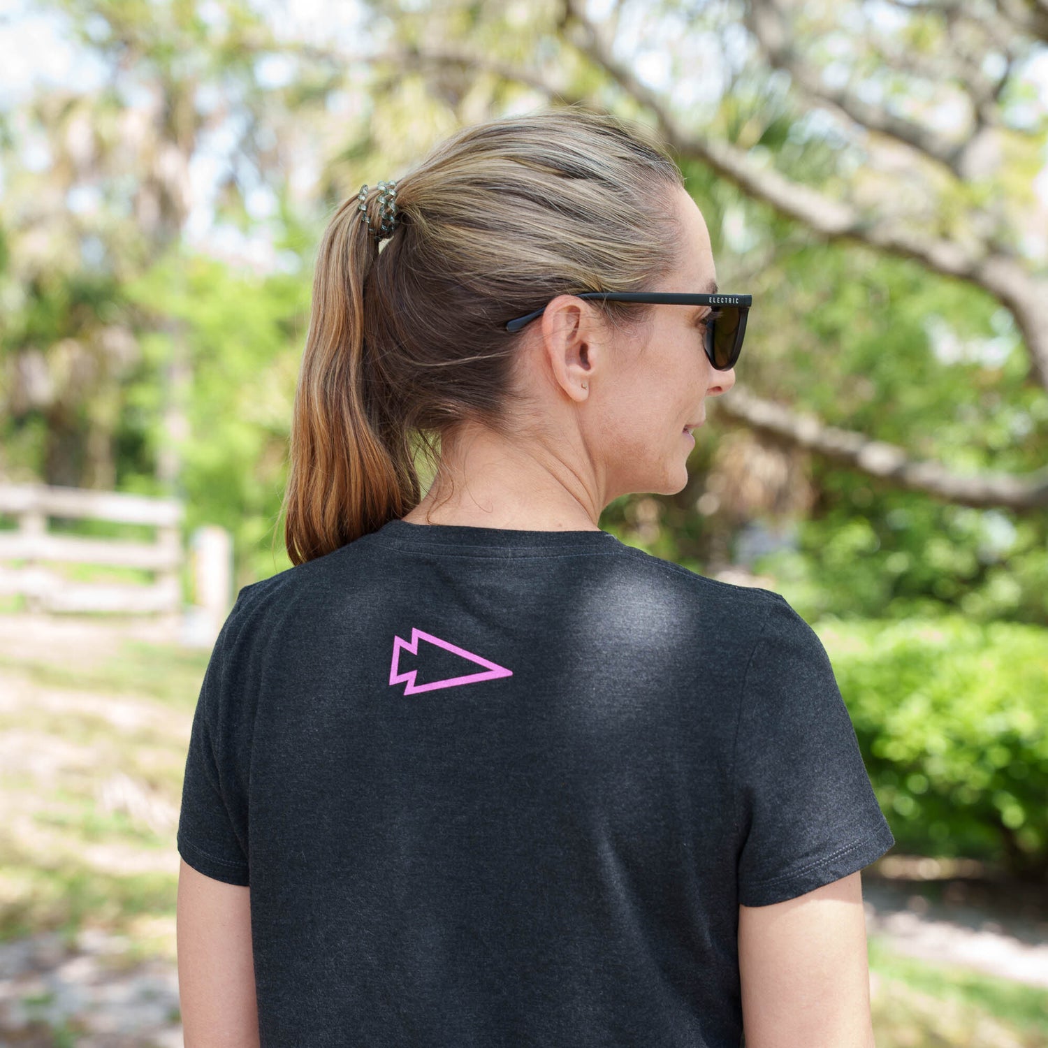 A woman with a ponytail and sunglasses enjoys the outdoors wearing the GORUCK MOTHERUCKER Women's Tee - Tri-BlendX, a black t-shirt with a pink arrow design on the back, offering a flattering fit and athletic cut.