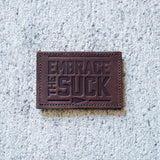 The Patch - Embrace The Suck features embossed lettering on brown leather, shown on concrete—an eye-catching addition to any Velcro patch collection.