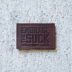 The Patch - Embrace The Suck features embossed lettering on brown leather, shown on concrete—an eye-catching addition to any Velcro patch collection.