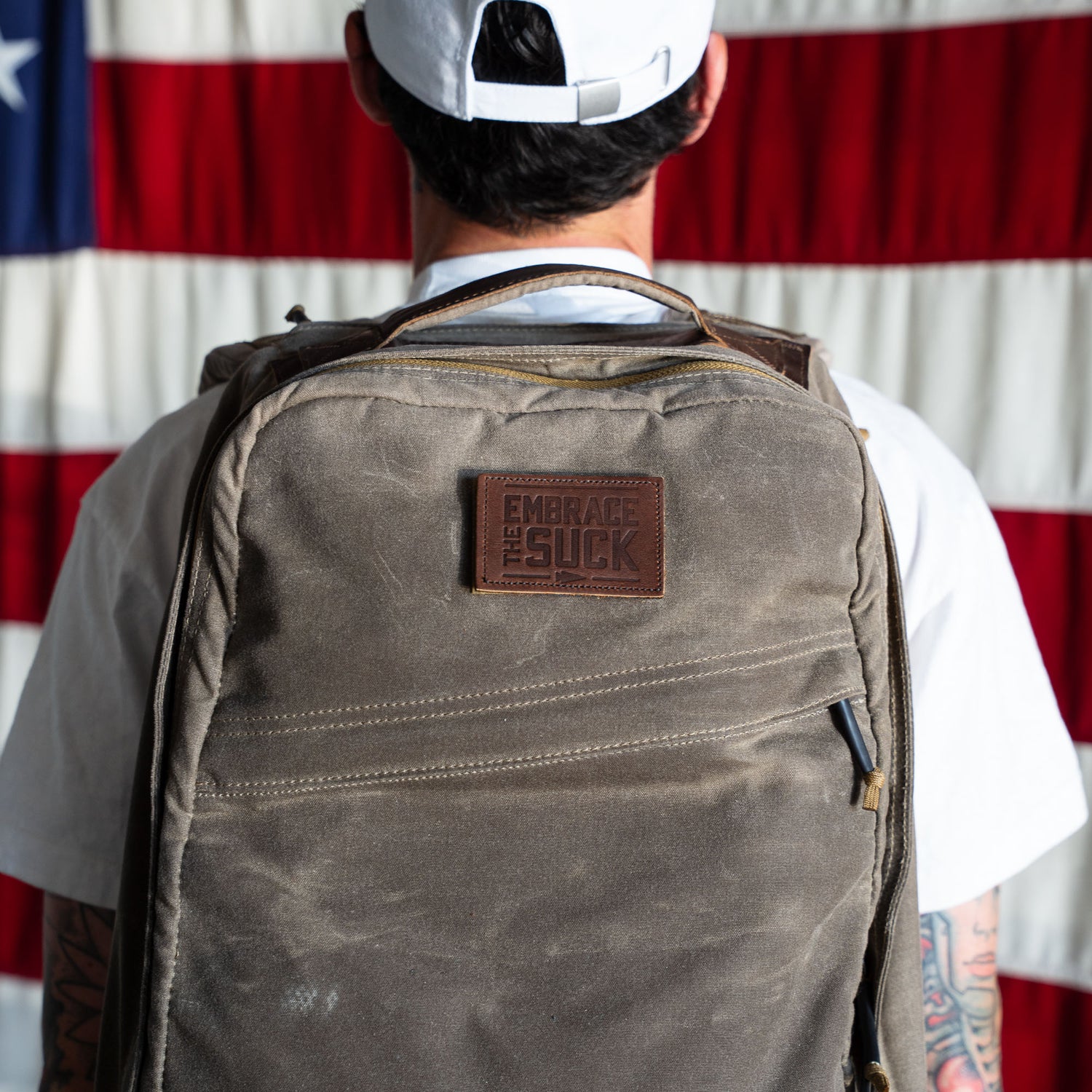 Man with a gray backpack displaying the Patch - Embrace The Suck stands in front of an American flag.