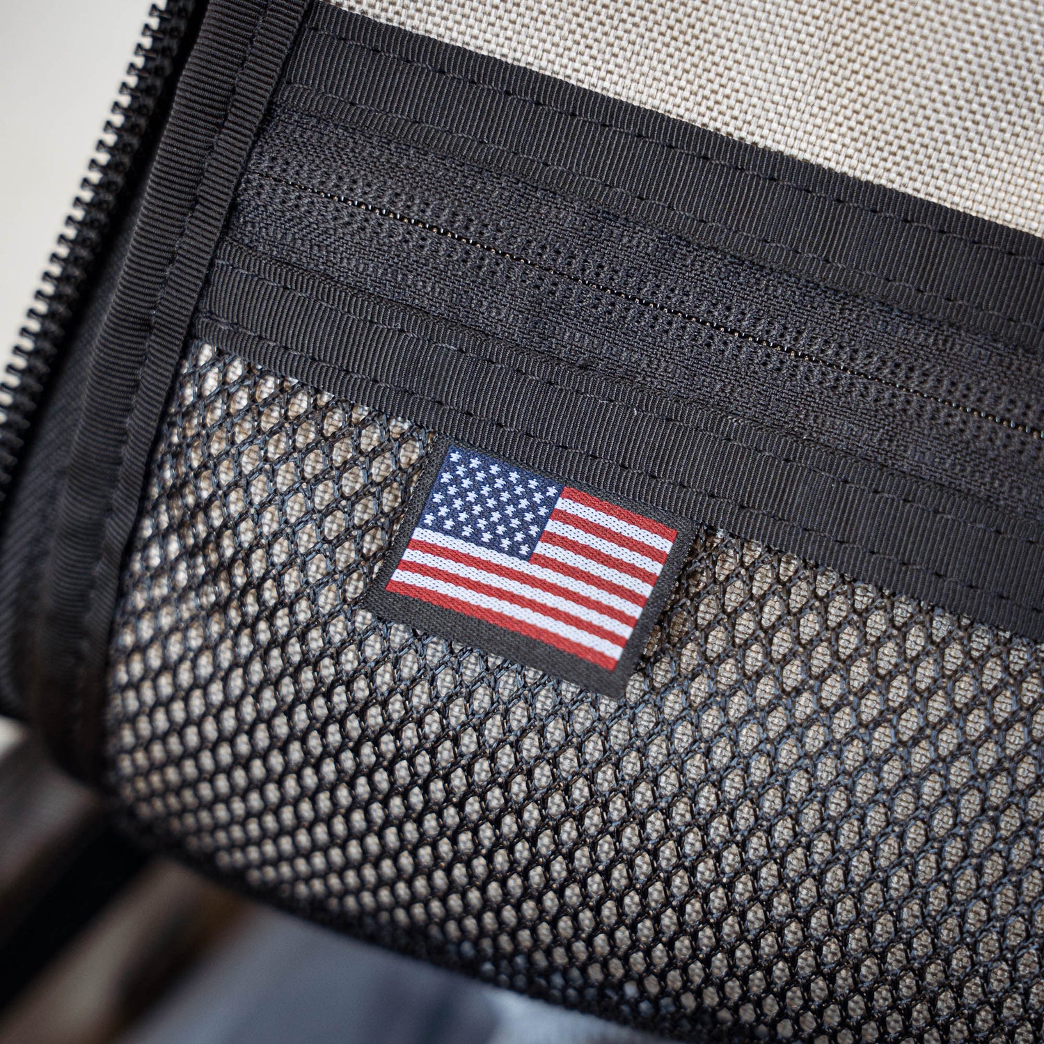 A small American flag patch is sewn onto black mesh fabric with a visible zipper, echoing details found on the GR1 backpack.