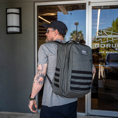 A man wearing sunglasses and carrying a GR2 travel backpack stands outside a building with glass doors displaying the "GORUCK" logo.