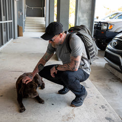 A man with tattoos and a GR2 rucksack kneels to pet a brown dog on a concrete sidewalk near parked cars.