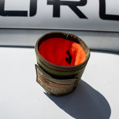 The Ballistic Beer Jacket, a green fabric koozie with an orange lining, rests on a white surface in sunlight.