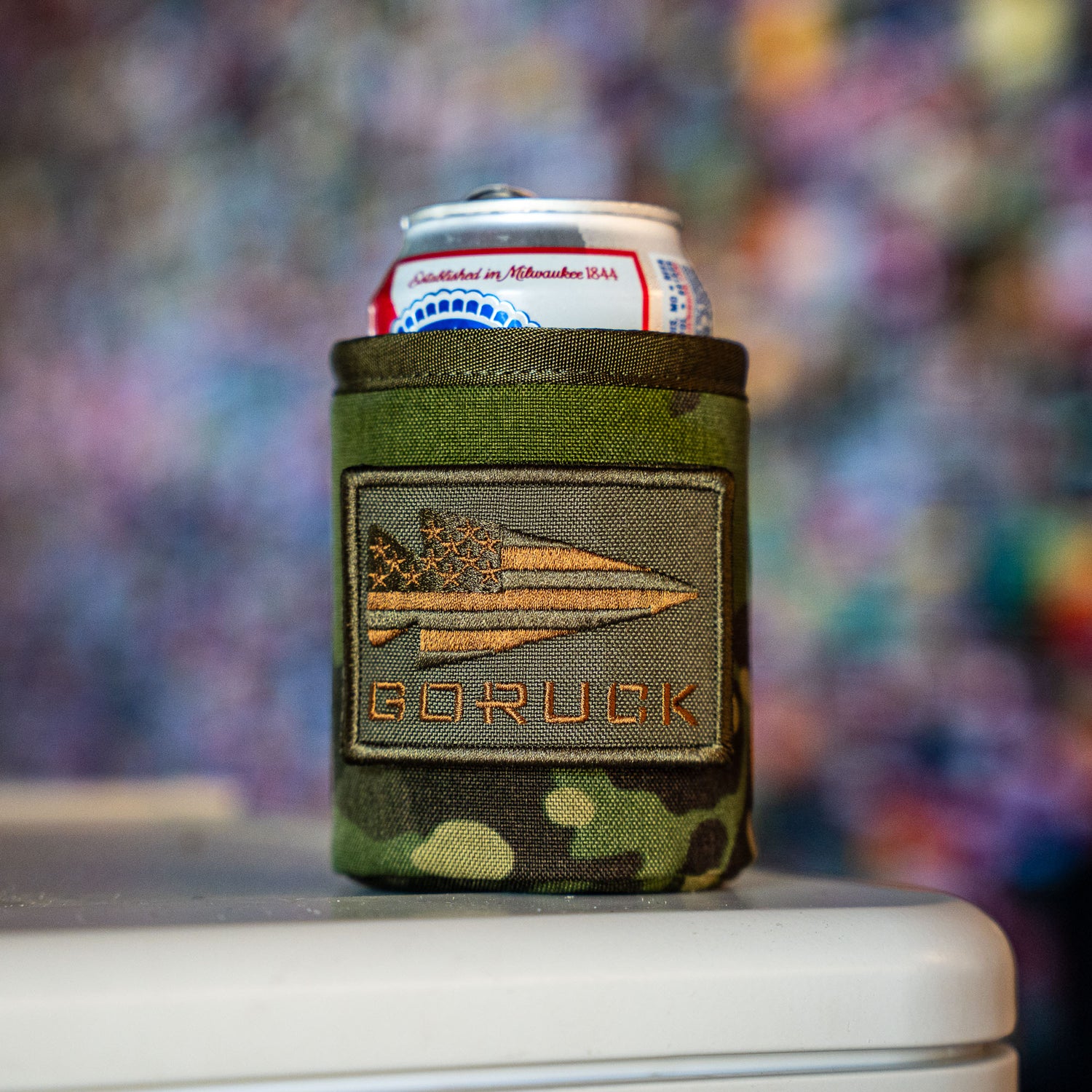 A can of Pabst Blue Ribbon beer in a Ballistic Beer Jacket by GORUCK, featuring camouflage material and an arrow and flag patch.