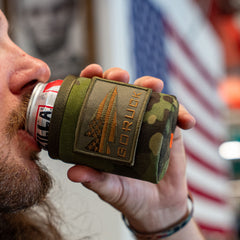 A person drinks from a can in a camo Ballistic Beer Jacket, with a blurred American flag in the background—perfect for keeping your drink cold, just like your favorite collapsible cooler.