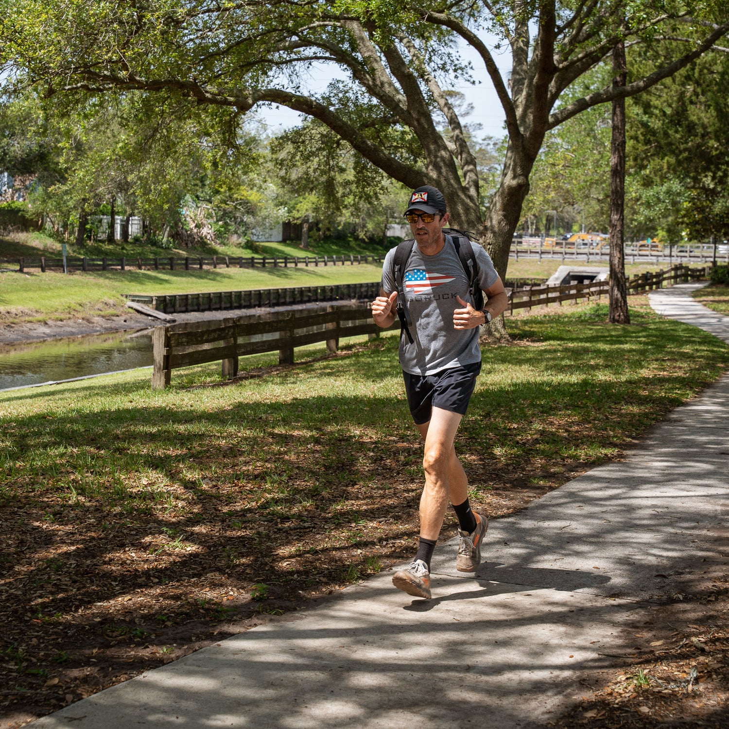 A man jogs on a sunny park path lined with trees and grass, wearing Men’s Training Shorts - ToughStretch, along with a backpack, cap, and sunglasses.