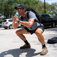 A man wearing the Mackall 45 Fog sunglasses and a backpack does a squat outdoors in a parking lot on a sunny day.