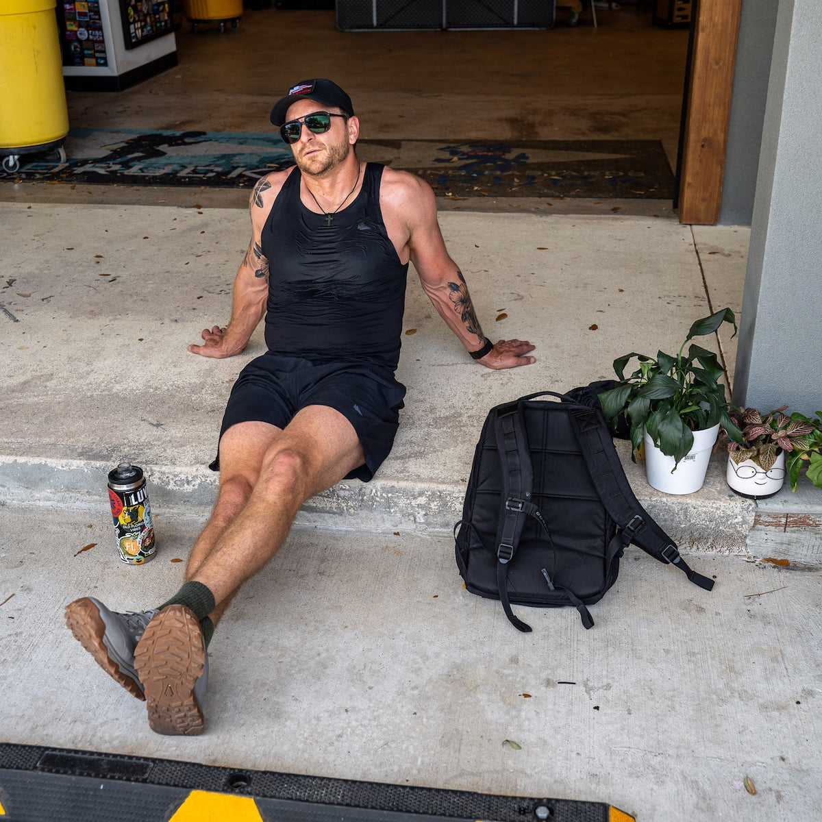 Man in black Men’s Performance Tank - ToughMesh and shorts sits on concrete, sunglasses on, with a backpack, drink, and potted plant nearby.
