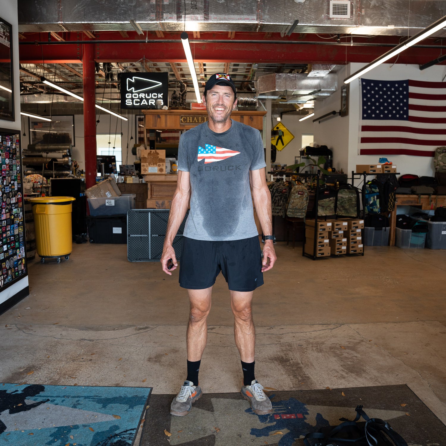 A man in athletic wear smiles in a warehouse with an American flag and gear behind him, wearing Men’s Training Shorts - ToughStretch, featuring sweat-wicking performance fabric.