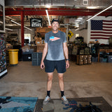 A man in athletic wear smiles in a warehouse with an American flag and gear behind him, wearing Men’s Training Shorts - ToughStretch, featuring sweat-wicking performance fabric.