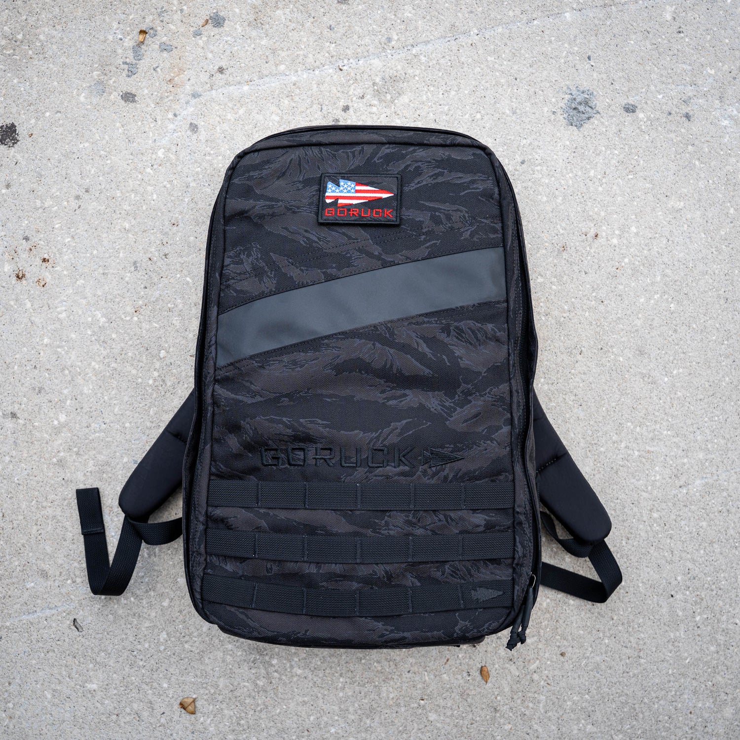 The black GORUCK Rucker 5.0 with a US flag patch, built for rucking with lumbar support, rests on rough, light gray concrete.