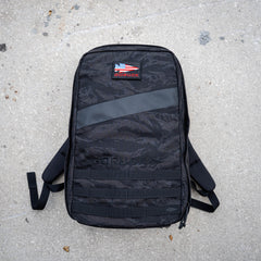 The black GORUCK Rucker 5.0 with a US flag patch, built for rucking with lumbar support, rests on rough, light gray concrete.