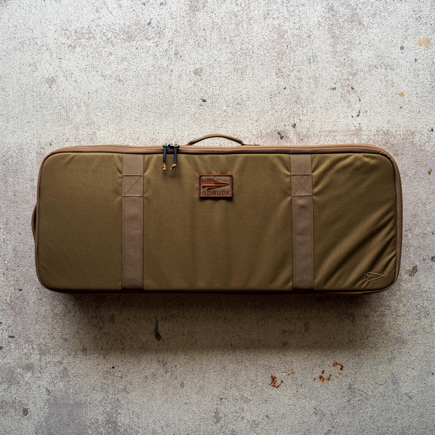 A rectangular brown Rifle Case with handles rests on a speckled concrete floor.