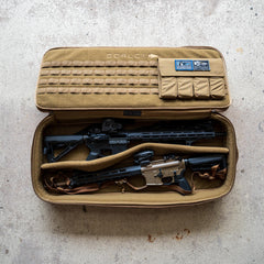 A Rifle Case in tan, adorned with patches, lies open on a concrete floor, displaying two rifles inside.