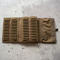 An Ammo Book in tan fabric lies open on a concrete surface, displaying multiple rifle cartridges secured in elastic loops.
