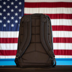 The GR1 Shooter 26L Flash Buffalo Fuzz - Built by Scars backpack is displayed upright with an American flag in the background, highlighting its handmade construction in Jax Beach, Florida.