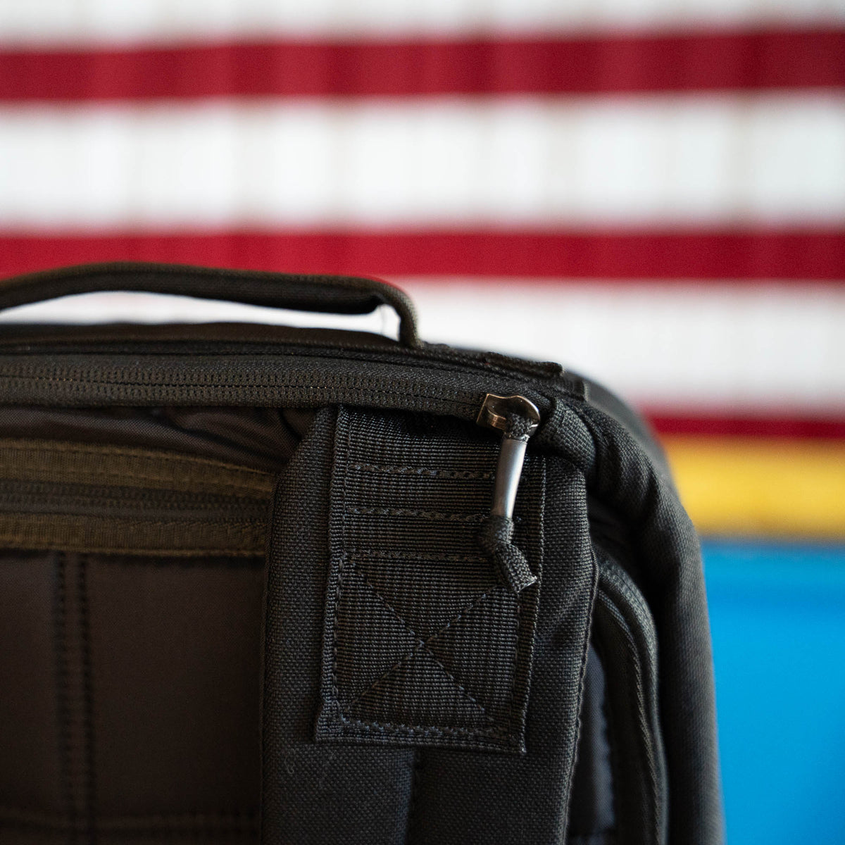 Close-up of a black bag's zipper and handle, the handmade GR1 Shooter 26L Flash Buffalo Fuzz - Built by Scars, with a blurred American flag in the background. This limited edition piece captures the spirit of craftsmanship from Jax Beach, Florida.