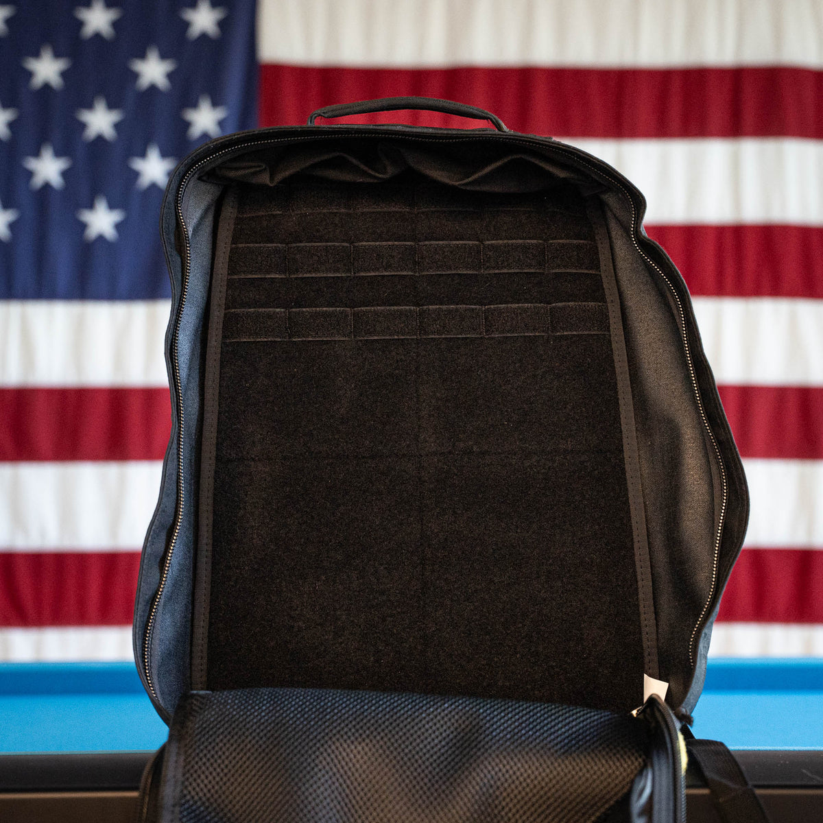 An open empty black GR1 Shooter 26L Flash Buffalo Fuzz - Built by Scars, a limited edition handmade backpack from Jax Beach, Florida, sits on a table with a USA flag hanging in the background.