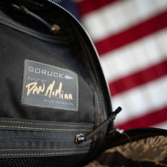 Tiger Stripe Robic Bullet – Built by Scars label with signature, partially open zipper, and a blurred American flag in the background—highlighting this limited edition, handmade rucksack inspired by Jax Beach, Florida.