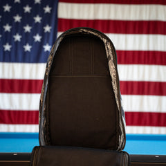 A black, limited edition Tiger Stripe Robic Bullet - Built by Scars backpack stands open on a table, with a U.S. flag hanging in the background—a handmade rucksack inspired by the coastal spirit of Jax Beach, Florida.