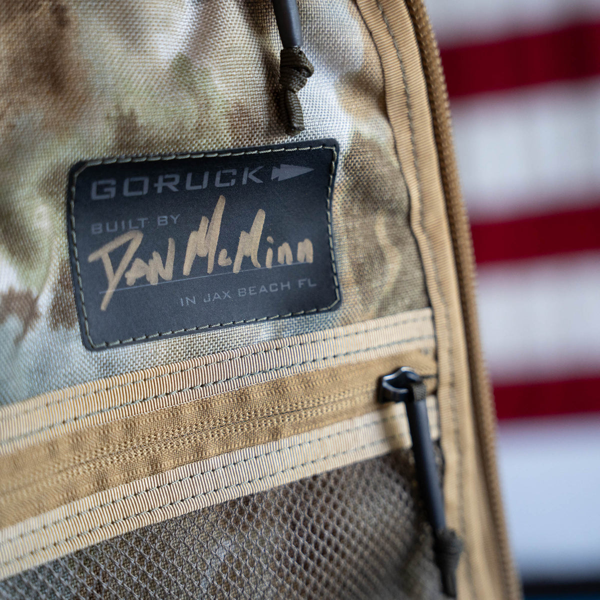 Close-up of a handmade Arid Bullet - Built by Scars ruck label with a signature and an American flag blurred in the background, crafted in Jax Beach, Florida as part of a limited edition release.