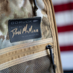 Close-up of a handmade Arid Bullet - Built by Scars ruck label with a signature and an American flag blurred in the background, crafted in Jax Beach, Florida as part of a limited edition release.