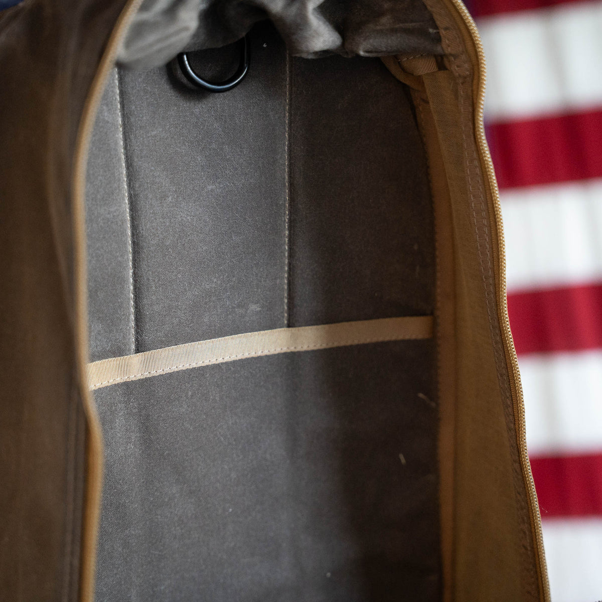 Close-up of an empty, limited edition handmade Waxed Bullet ruck with a tan interior pocket and a zipper, with part of a red and white striped object visible behind it.