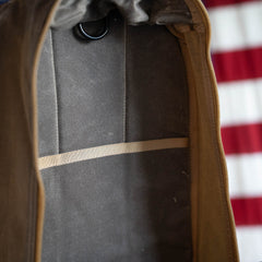 Close-up of an empty, limited edition handmade Waxed Bullet ruck with a tan interior pocket and a zipper, with part of a red and white striped object visible behind it.