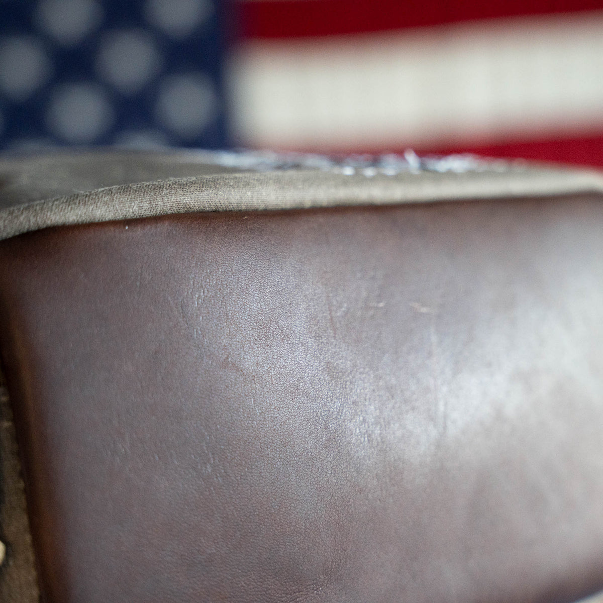 Close-up of the Waxed Bullet, a brown leather, handmade ruck with a blurred American flag in the background, capturing the spirit of Jax Beach, Florida. This limited edition piece combines expert craftsmanship and local pride in every detail.