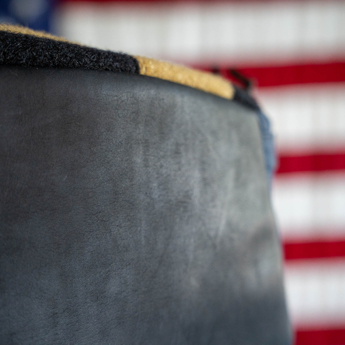 Close-up of the Checkered GR1 21L - Built by Scars leather surface, with part of an American flag visible in the blurred background, highlighting the expert craftsmanship and unique design inspired by Jax Beach, Florida.