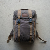 The M26 PREORDER // Q4 2026 ETA is a brown canvas backpack with tan straps and a leather patch, shown on a light concrete surface and featuring a bombproof laptop compartment for extra protection.