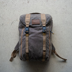 The M26 PREORDER // Q4 2026 ETA is a brown canvas backpack with tan straps and a leather patch, shown on a light concrete surface and featuring a bombproof laptop compartment for extra protection.