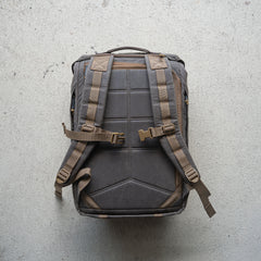 The M26 PREORDER // Q4 2026 ETA backpack, featuring padded straps, buckles, and a bombproof laptop compartment, is displayed flat on a textured light-colored surface.
