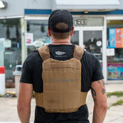 A man in a black cap and shirt wears the Achilles | Men's Weighted Vest in tan, standing outside a store with his back to the camera.