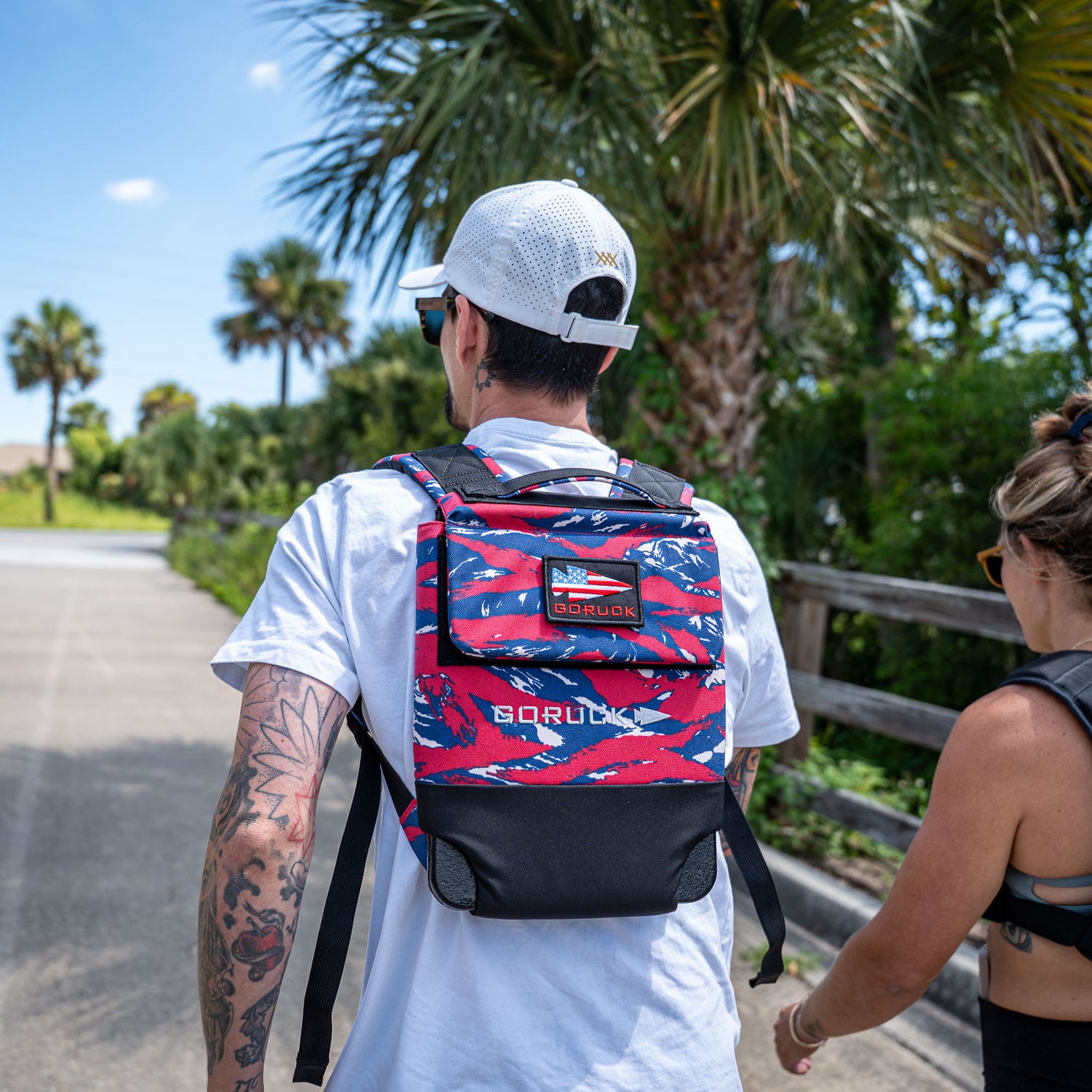 A man walks outdoors with a woman on a sunny day, palm trees in the background, wearing a Ruck Plate Carrier 3.0 backpack featuring ergonomic lumbar support in red, white, and blue.